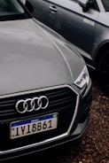A close-up of the front of an Audi car with a Brazilian license plate, parked on a gravel surface. The Audi emblem is prominently visible on the front grille. The car is positioned next to another vehicle, which is partially visible in the background.