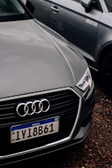 A close-up of the front of an Audi car with a Brazilian license plate, parked on a gravel surface. The Audi emblem is prominently visible on the front grille. The car is positioned next to another vehicle, which is partially visible in the background.