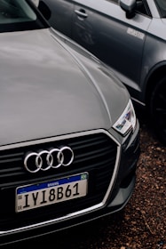 A close-up of the front of an Audi car with a Brazilian license plate, parked on a gravel surface. The Audi emblem is prominently visible on the front grille. The car is positioned next to another vehicle, which is partially visible in the background.
