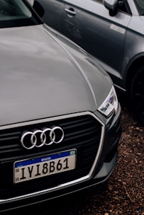 A close-up of the front of an Audi car with a Brazilian license plate, parked on a gravel surface. The Audi emblem is prominently visible on the front grille. The car is positioned next to another vehicle, which is partially visible in the background.