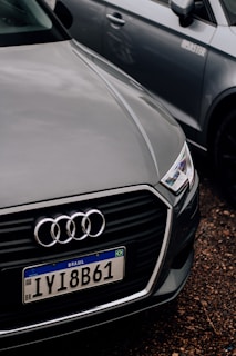 A close-up of the front of an Audi car with a Brazilian license plate, parked on a gravel surface. The Audi emblem is prominently visible on the front grille. The car is positioned next to another vehicle, which is partially visible in the background.