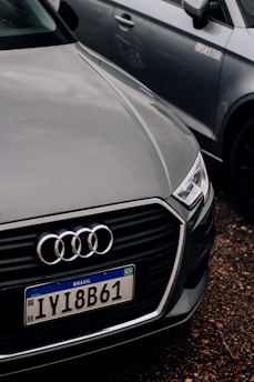 A close-up of the front of an Audi car with a Brazilian license plate, parked on a gravel surface. The Audi emblem is prominently visible on the front grille. The car is positioned next to another vehicle, which is partially visible in the background.