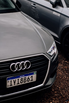 A close-up of the front of an Audi car with a Brazilian license plate, parked on a gravel surface. The Audi emblem is prominently visible on the front grille. The car is positioned next to another vehicle, which is partially visible in the background.