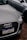 A close-up of the front of an Audi car with a Brazilian license plate, parked on a gravel surface. The Audi emblem is prominently visible on the front grille. The car is positioned next to another vehicle, which is partially visible in the background.
