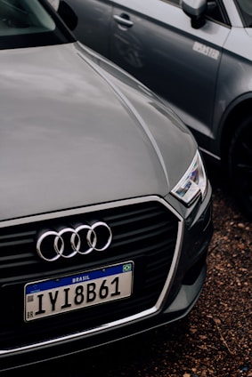 A close-up of the front of an Audi car with a Brazilian license plate, parked on a gravel surface. The Audi emblem is prominently visible on the front grille. The car is positioned next to another vehicle, which is partially visible in the background.