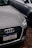 A close-up of the front of an Audi car with a Brazilian license plate, parked on a gravel surface. The Audi emblem is prominently visible on the front grille. The car is positioned next to another vehicle, which is partially visible in the background.