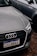 A close-up of the front of an Audi car with a Brazilian license plate, parked on a gravel surface. The Audi emblem is prominently visible on the front grille. The car is positioned next to another vehicle, which is partially visible in the background.