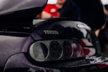 A close-up view of the rear section of a black car, featuring the Toyota logo prominently displayed. The car surface is covered with raindrops, suggesting a rainy or damp environment. The background is blurred with some indistinct people and a hint of red clothing.
