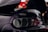 A close-up view of the rear section of a black car, featuring the Toyota logo prominently displayed. The car surface is covered with raindrops, suggesting a rainy or damp environment. The background is blurred with some indistinct people and a hint of red clothing.