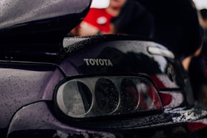 A close-up view of the rear section of a black car, featuring the Toyota logo prominently displayed. The car surface is covered with raindrops, suggesting a rainy or damp environment. The background is blurred with some indistinct people and a hint of red clothing.