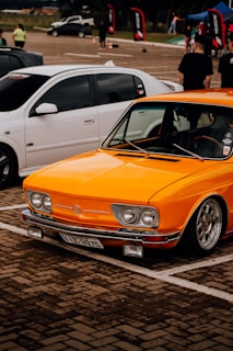 An orange vintage car with chrome details is parked next to a white car in a paved outdoor area, possibly during a car show. Several people are visible in the background, along with flags and a tent, suggesting an event atmosphere.