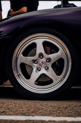 A close-up of a polished alloy car wheel with a visible HRE logo in the center. The wheel is part of a polished tire attached to a dark-colored vehicle. The background is blurred with indistinct shapes, possibly people or objects near the car.