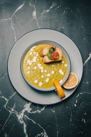 A bowl of green soup with white drizzle and two pieces of bread garnished with herbs and flowers on top, situated on a gray plate with a slice of lemon on the side. The background consists of a dark marble surface with white veining.