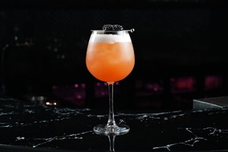 A vibrant orange cocktail served in a wide glass with a foamy top, garnished with blackberries on a skewer. The drink is set against a dark, blurred background, creating a sophisticated and elegant presentation.