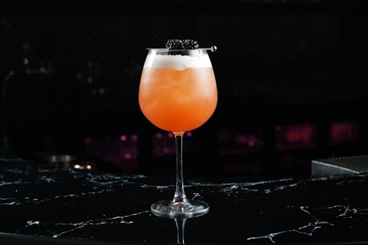A vibrant orange cocktail served in a wide glass with a foamy top, garnished with blackberries on a skewer. The drink is set against a dark, blurred background, creating a sophisticated and elegant presentation.