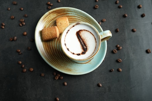 A cup of frothy coffee with latte art, accompanied by two shortbread cookies, sits on a saucer. Coffee beans are scattered across the dark surface around the cup.