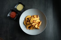 A plate of golden brown potato wedges is served in a light-colored bowl. Next to the bowl, there are two small square dishes, one containing a creamy dip and the other filled with ketchup. The surfaces are set against a dark, textured background.