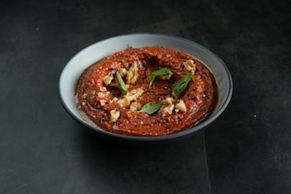 A bowl filled with a rich, red-brown dip topped with chopped nuts and green leaves. The dip has a smooth texture and is garnished with speckles of black seeds.