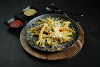 A wooden platter holds a plate of seasoned french fries topped with melted cheese and garnished with herbs. Two small black bowls are placed on the side, one containing ketchup and the other a yellow sauce. Two forks are positioned behind the plate, resting on the table.