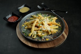 A wooden platter holds a plate of seasoned french fries topped with melted cheese and garnished with herbs. Two small black bowls are placed on the side, one containing ketchup and the other a yellow sauce. Two forks are positioned behind the plate, resting on the table.