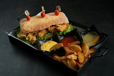 A ciabatta sandwich filled with crispy fried chicken, lettuce, and tomato is garnished with three cherry tomatoes skewered on top. It is served on a tray alongside crispy potato wedges and three dips containing yellow mustard, red ketchup, and a creamy green sauce.
