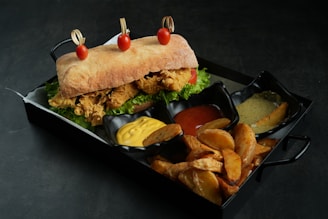 A ciabatta sandwich filled with crispy fried chicken, lettuce, and tomato is garnished with three cherry tomatoes skewered on top. It is served on a tray alongside crispy potato wedges and three dips containing yellow mustard, red ketchup, and a creamy green sauce.