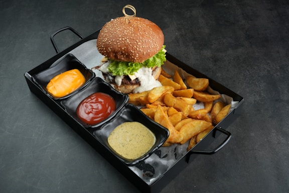 A gourmet burger topped with lettuce is placed on a tray accompanied by golden potato wedges and three small dishes filled with orange cheese sauce, red ketchup, and a creamy mustard sauce.