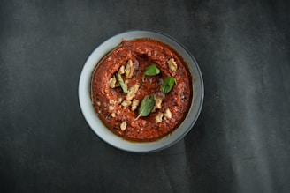 A bowl containing a thick red sauce or dip garnished with pieces of walnuts and fresh green basil leaves is placed on a dark surface, providing a contrasting backdrop to the vibrant dish.