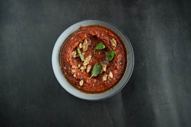 A bowl containing a thick red sauce or dip garnished with pieces of walnuts and fresh green basil leaves is placed on a dark surface, providing a contrasting backdrop to the vibrant dish.