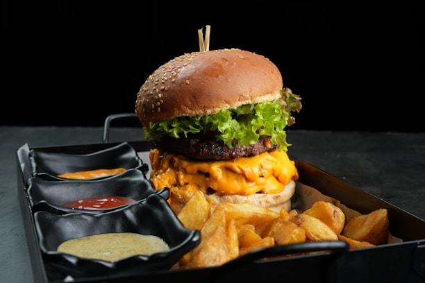 A gourmet burger with a sesame seed bun, melted cheese, and fresh green lettuce is presented on a black tray. Accompanying the burger are crispy potato wedges and three small black dishes containing different sauces: one with mustard, another with ketchup, and a third with a creamy sauce.