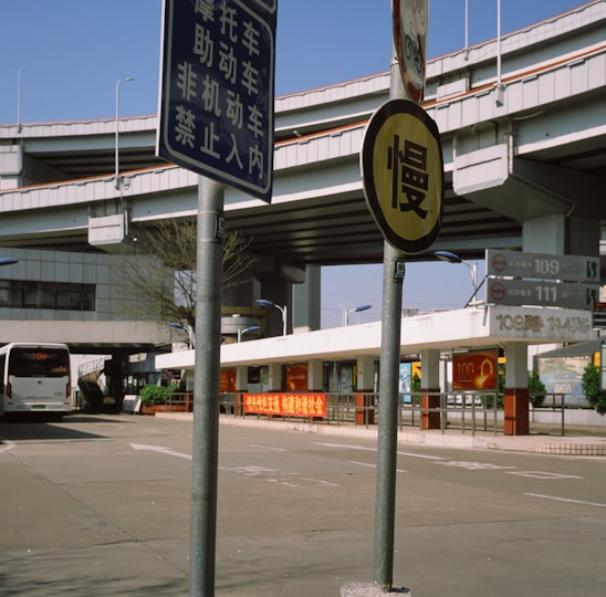 An urban landscape featuring a large multi-layered highway interchange with multiple road signs and a bus station. The foreground displays Chinese characters on the signs, and a bus can be seen approaching the station. The environment is well-lit with clear skies, and some greenery is visible.