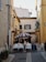 A narrow, charming street features outdoor dining with tables set under a large brown and white striped umbrella. The street is flanked by colorful buildings with shuttered windows and plants adorning balconies. A cozy, intimate atmosphere is created by the stone pavement and the quaint arrangement of the café.