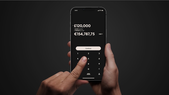 A person holding a smartphone with both hands against a dark background. The screen displays a calculator-like interface with a financial transaction and currency conversion in pounds and euros, along with fee information. A finger is pressing a button labeled 'Continue'.