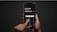 A person holding a smartphone with both hands against a dark background. The screen displays a calculator-like interface with a financial transaction and currency conversion in pounds and euros, along with fee information. A finger is pressing a button labeled 'Continue'.