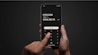 A person holding a smartphone with both hands against a dark background. The screen displays a calculator-like interface with a financial transaction and currency conversion in pounds and euros, along with fee information. A finger is pressing a button labeled 'Continue'.
