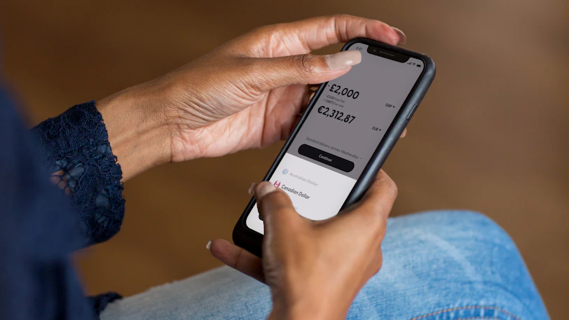 A traveler checking currency exchange rates on a smartphone at an airport lounge with a lilac-themed background.