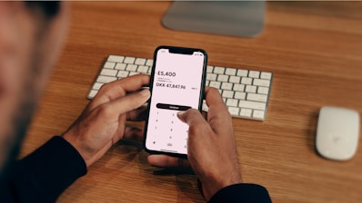 A person is holding a smartphone displaying a currency conversion app showing the exchange from GBP to DKK. The background contains a wooden desk with a keyboard and a computer mouse placed on it.