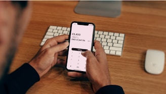 A person is holding a smartphone displaying a currency conversion app showing the exchange from GBP to DKK. The background contains a wooden desk with a keyboard and a computer mouse placed on it.