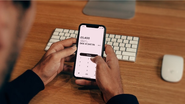A person is holding a smartphone displaying a currency conversion app showing the exchange from GBP to DKK. The background contains a wooden desk with a keyboard and a computer mouse placed on it.