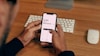 A person is holding a smartphone displaying a currency conversion app showing the exchange from GBP to DKK. The background contains a wooden desk with a keyboard and a computer mouse placed on it.