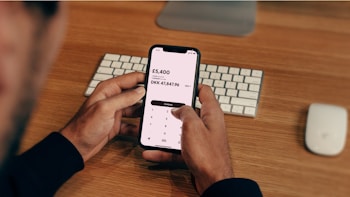 A person is holding a smartphone displaying a currency conversion app showing the exchange from GBP to DKK. The background contains a wooden desk with a keyboard and a computer mouse placed on it.