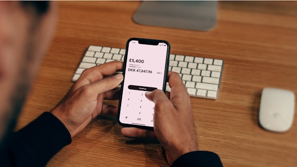 A person is holding a smartphone displaying a currency conversion app showing the exchange from GBP to DKK. The background contains a wooden desk with a keyboard and a computer mouse placed on it.