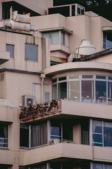 A section of a beige, multi-story building with large windows and balconies. The building has visible signs of wear and rust, particularly on the metal sign that appears to be corroded. Some air conditioning units are mounted on the walls, and the roof features various equipment and structures.