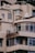 A section of a beige, multi-story building with large windows and balconies. The building has visible signs of wear and rust, particularly on the metal sign that appears to be corroded. Some air conditioning units are mounted on the walls, and the roof features various equipment and structures.