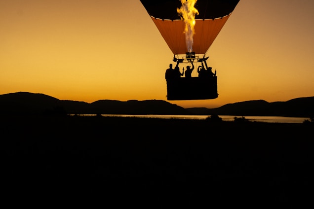 A hot air balloon silhouetted against a vibrant orange and yellow sunset sky. The balloon glows with flames as it rises, casting a stark contrast between the darkened landscape and the bright sky. The balloon basket holds several passengers, adding a sense of adventure and exploration.