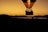 A hot air balloon silhouetted against a vibrant orange and yellow sunset sky. The balloon glows with flames as it rises, casting a stark contrast between the darkened landscape and the bright sky. The balloon basket holds several passengers, adding a sense of adventure and exploration.
