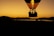 A hot air balloon silhouetted against a vibrant orange and yellow sunset sky. The balloon glows with flames as it rises, casting a stark contrast between the darkened landscape and the bright sky. The balloon basket holds several passengers, adding a sense of adventure and exploration.