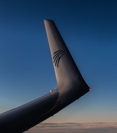 A close-up view of an airplane's vertical stabilizer or tail fin against a backdrop of a blue sky transitioning to a lighter gradient near the horizon. The tail fin has a logo featuring a stylized bird.