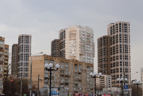 A series of tall, modern apartment buildings with multiple floors dominate the skyline, surrounded by more traditional, lower-rise buildings. The sky is overcast, suggesting a city scene during a cloudy day. Street lights and signs are visible in the foreground, indicating an urban environment.