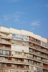 A multi-story residential building features numerous balconies with windows and air conditioning units. The structure is characterized by a mix of wood and glass elements, and sits against a clear blue sky.